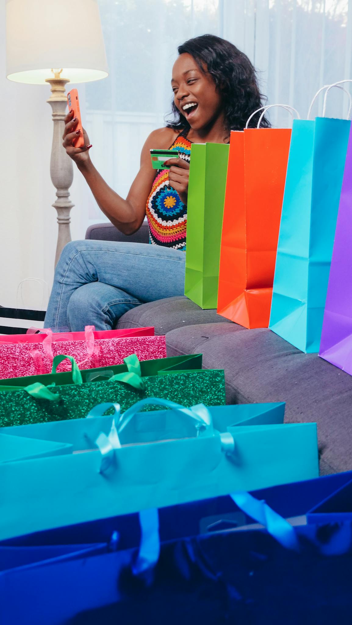 A woman enjoys online shopping with a smartphone, surrounded by colorful shopping bags indoors.